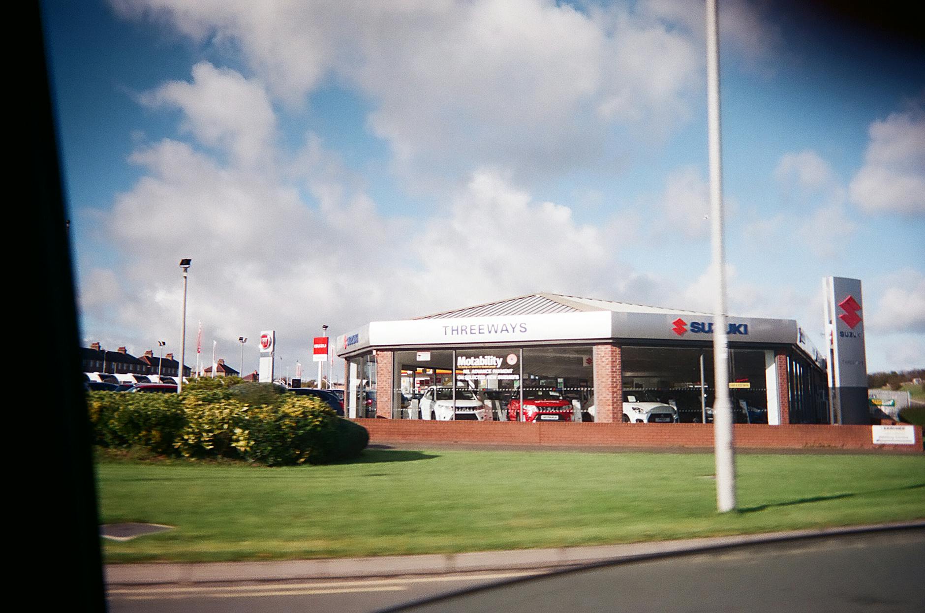 Ilustrasi: Exterior view of a car dealership featuring Suzuki automobiles under a bright sky.