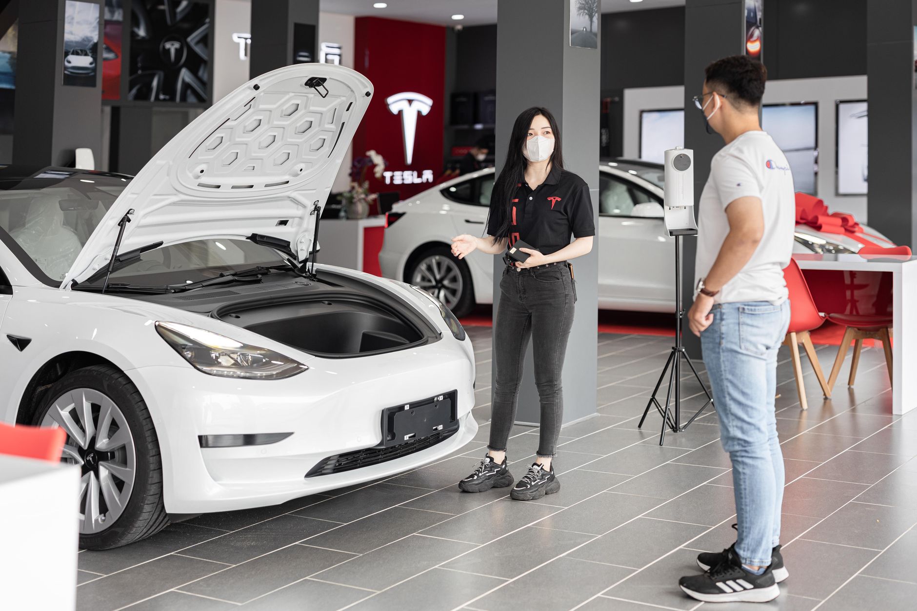 Ilustrasi: A customer talks with a sales representative about a Tesla Model 3 in a car dealership, showcasing the electric car's features.