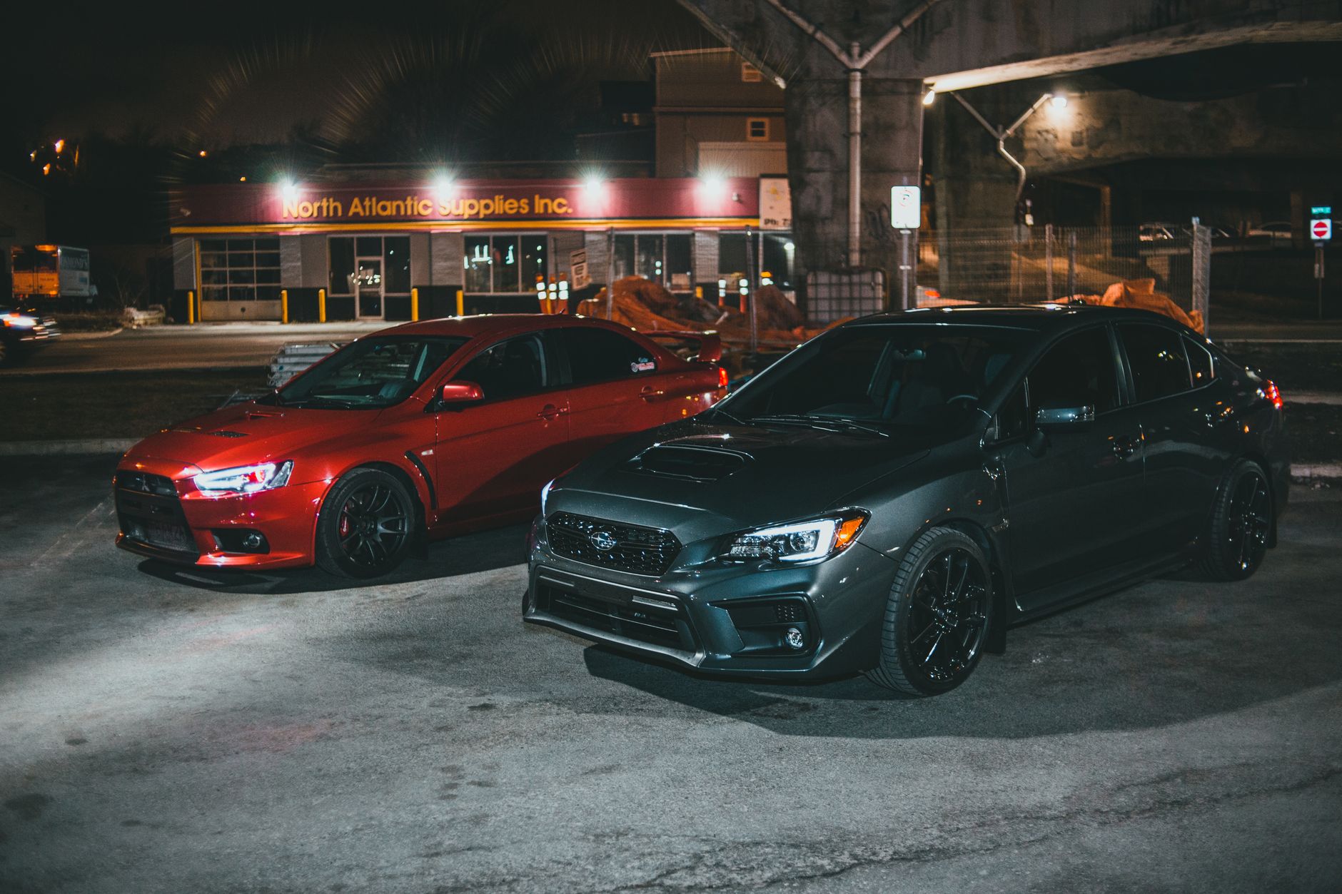 Ilustrasi: Two sport sedans, Mitsubishi Lancer EVO and Subaru WRX, parked under street lights malam hari.