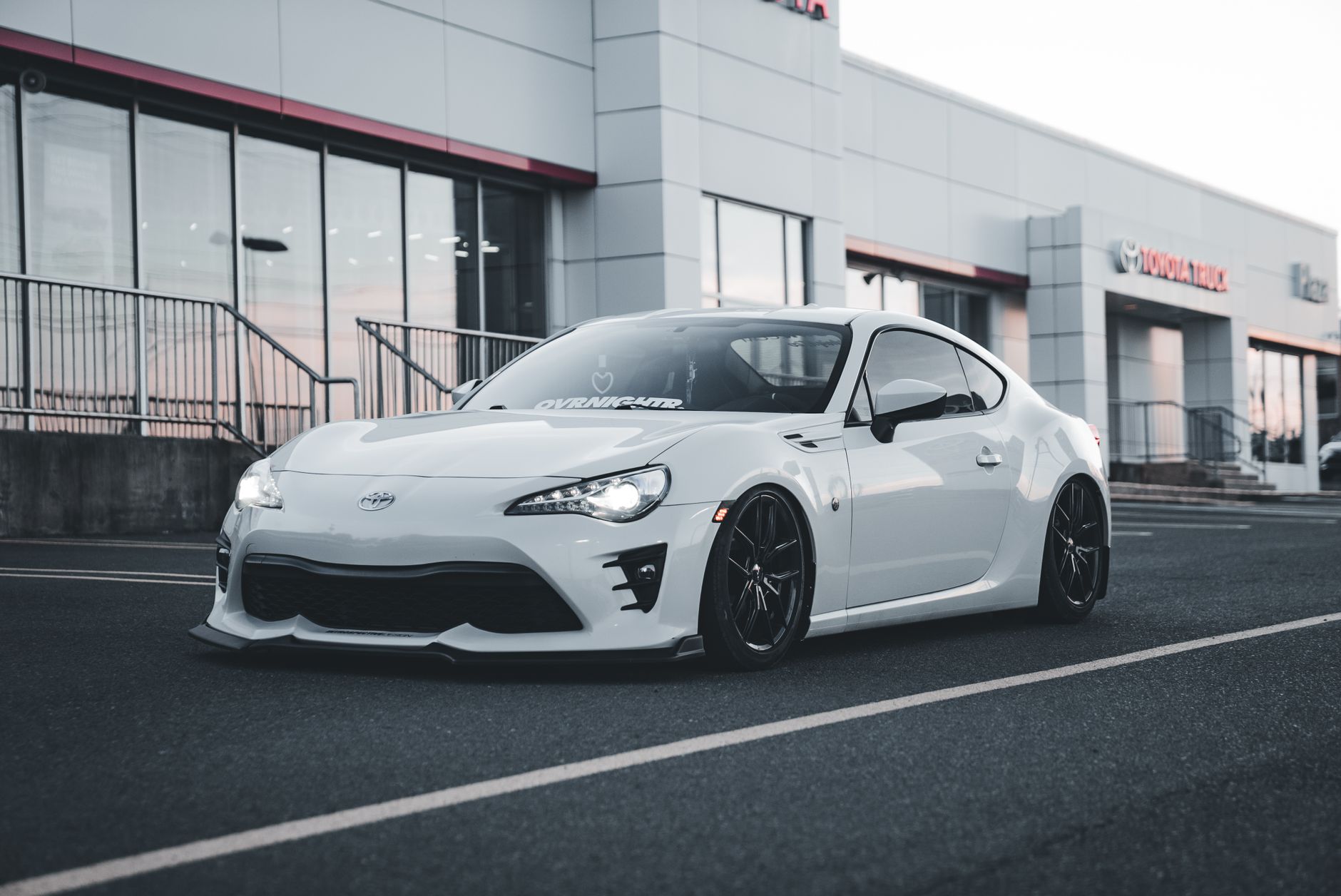 Ilustrasi: Stylish white sports car parked in front of Toyota dealership building at twilight.
