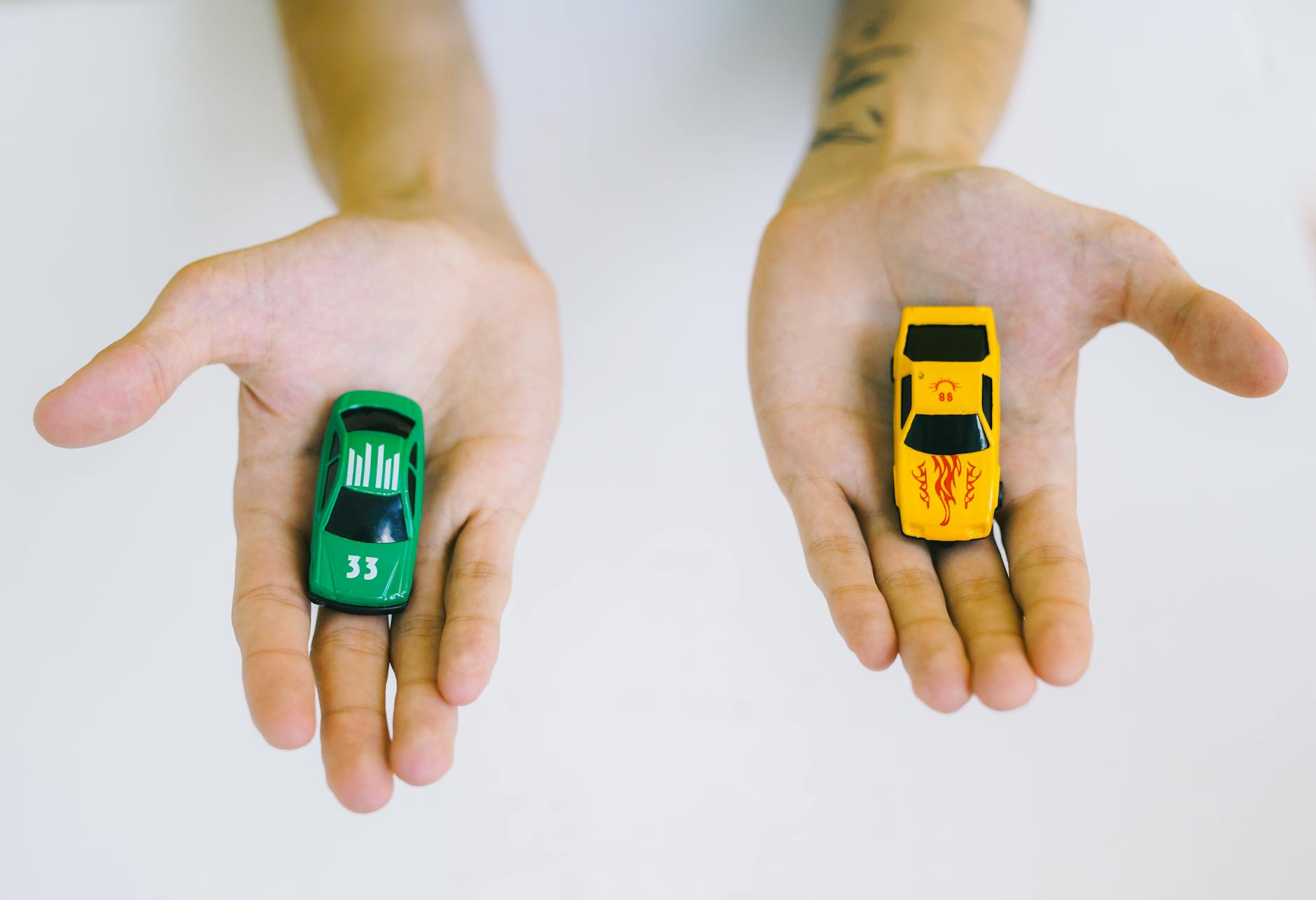 Ilustrasi: Pair of adult hands holding green and yellow toy cars on a white surface, symbolizing choice.