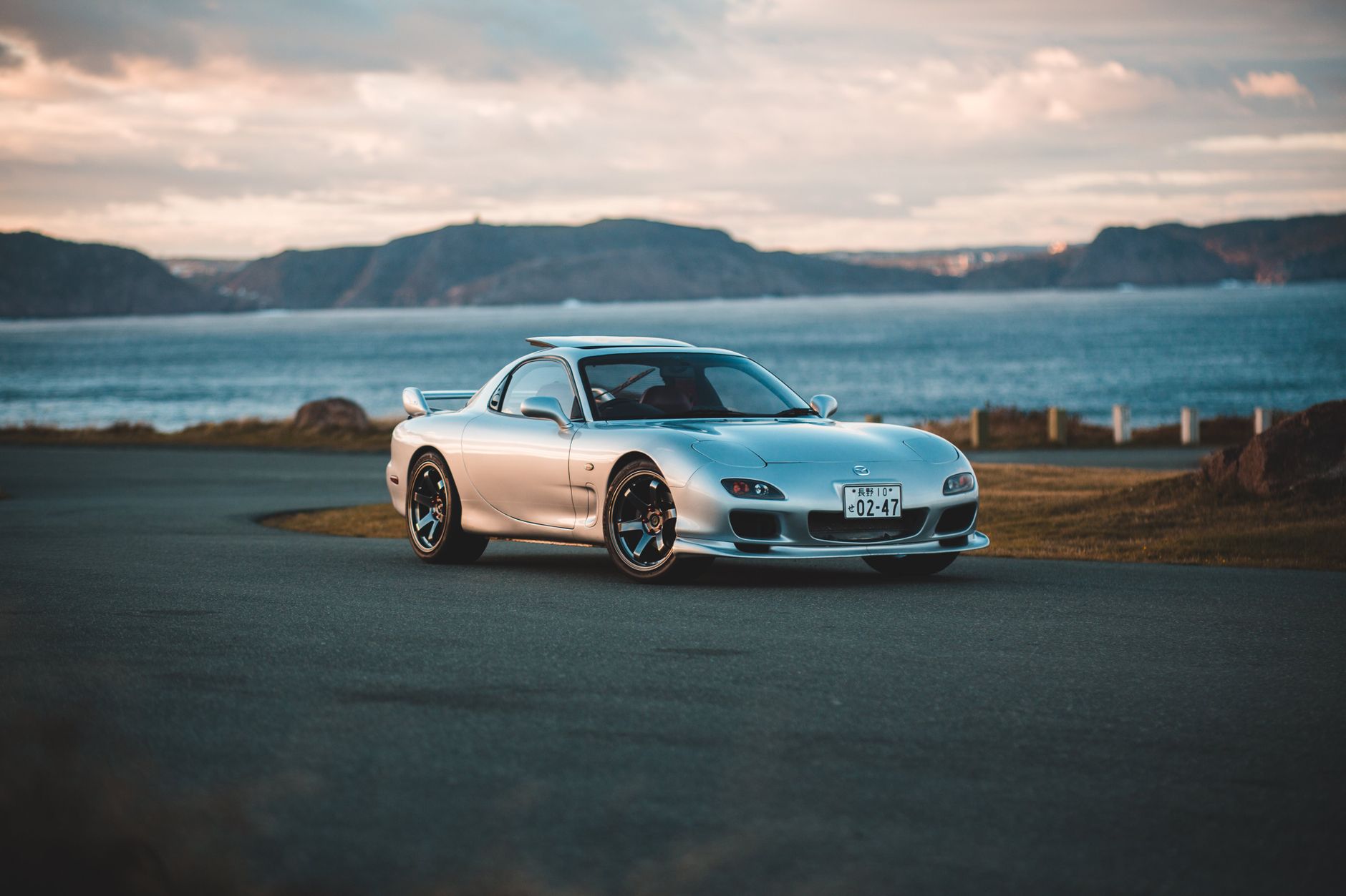 Ilustrasi: A sleek silver sports car parked by a scenic coastal road with mountains in the background.