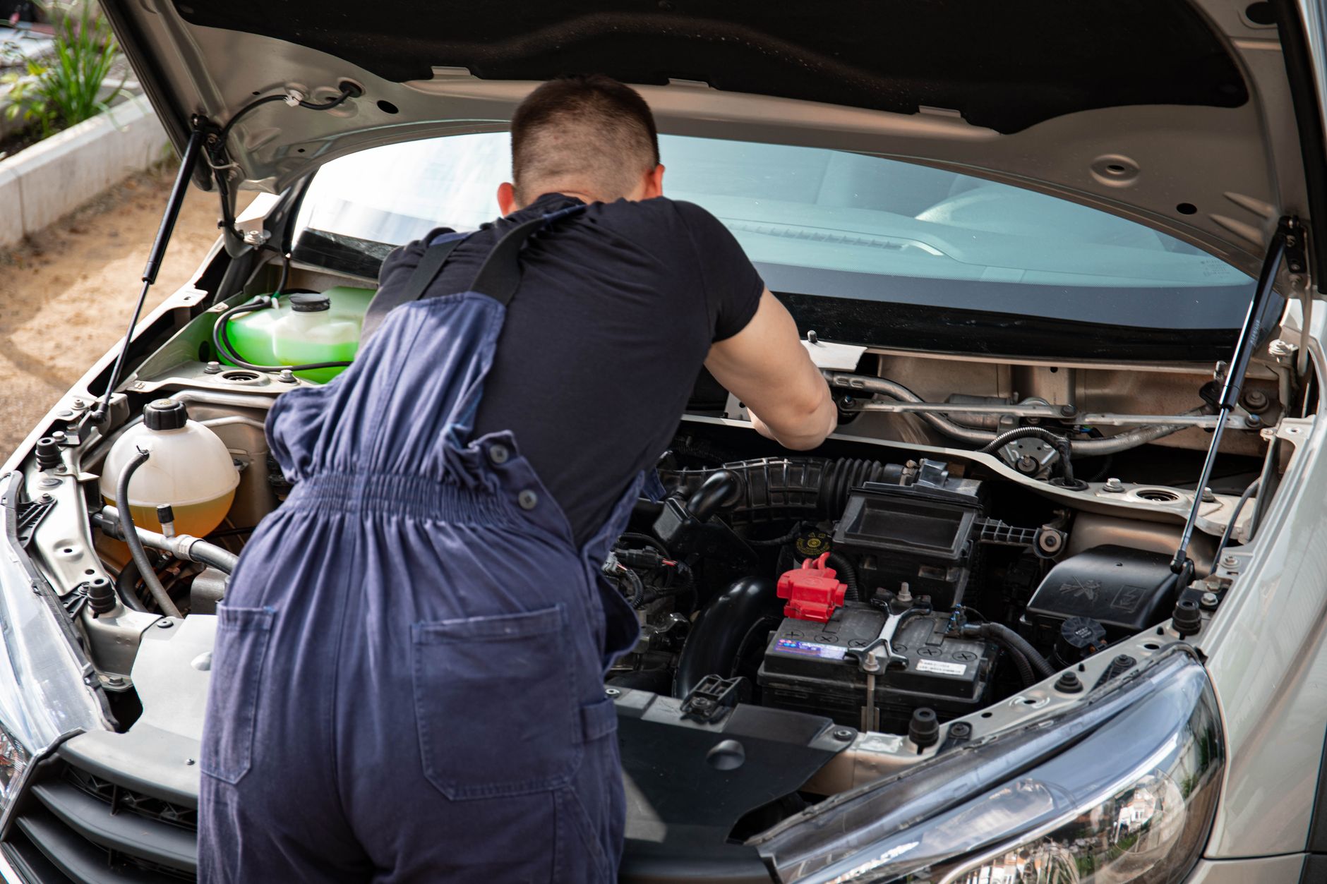 Ilustrasi: Mechanic in blue coveralls performing car engine maintenance outdoors.