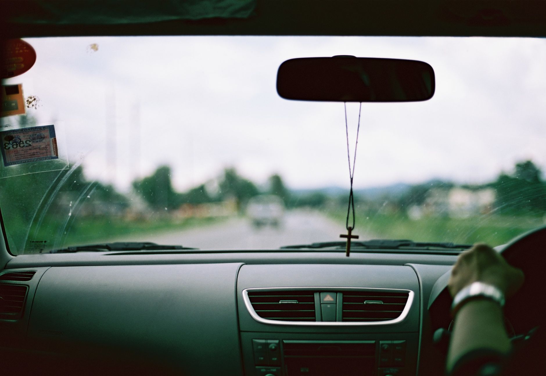 Ilustrasi: Interior view of a car dashboard with hanging cross on rear-view mirror, blurred road ahead.