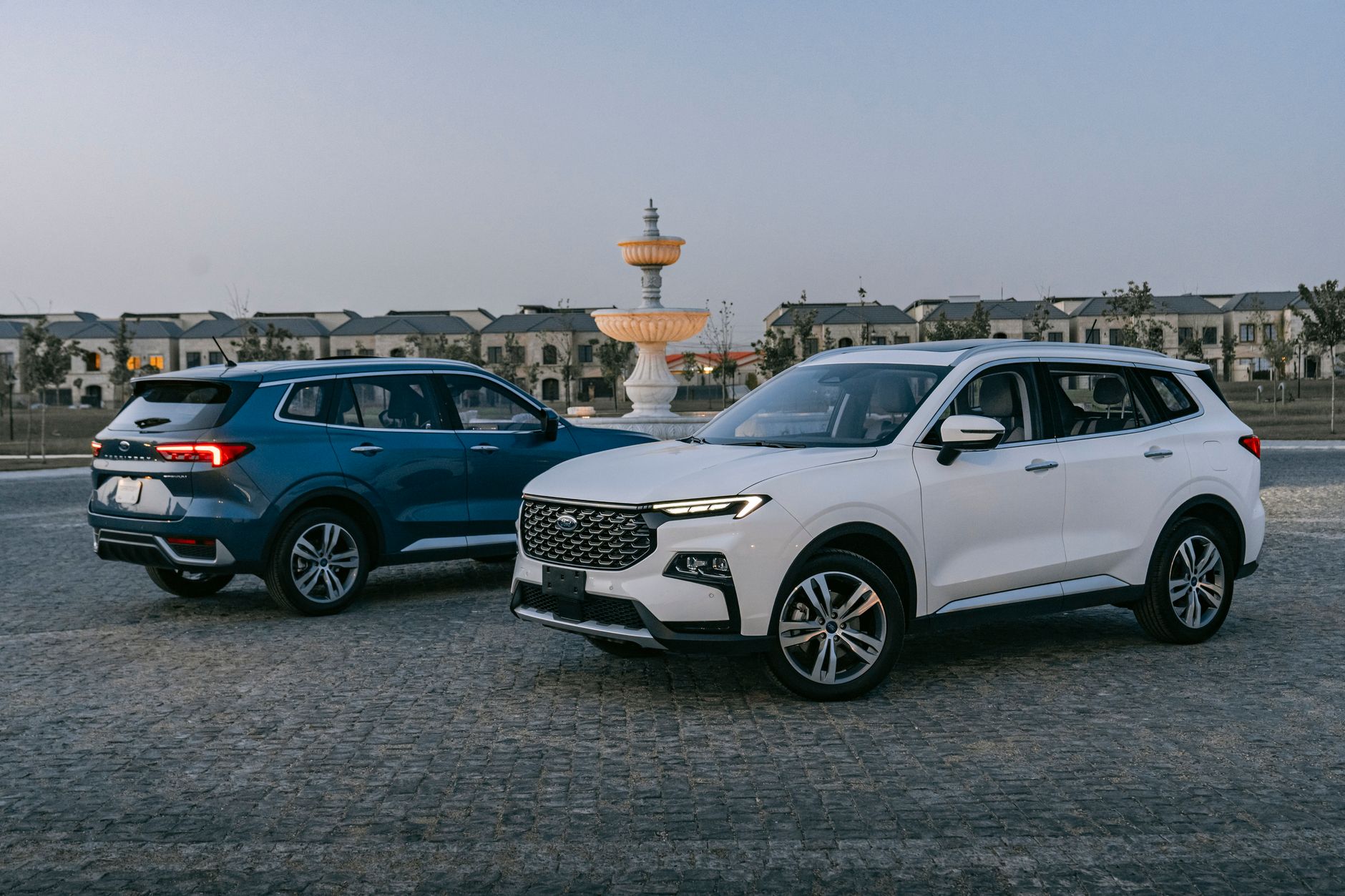 Ilustrasi: Two modern SUVs parked in a stylish plaza with a decorative fountain, showcasing contemporary automotive design.