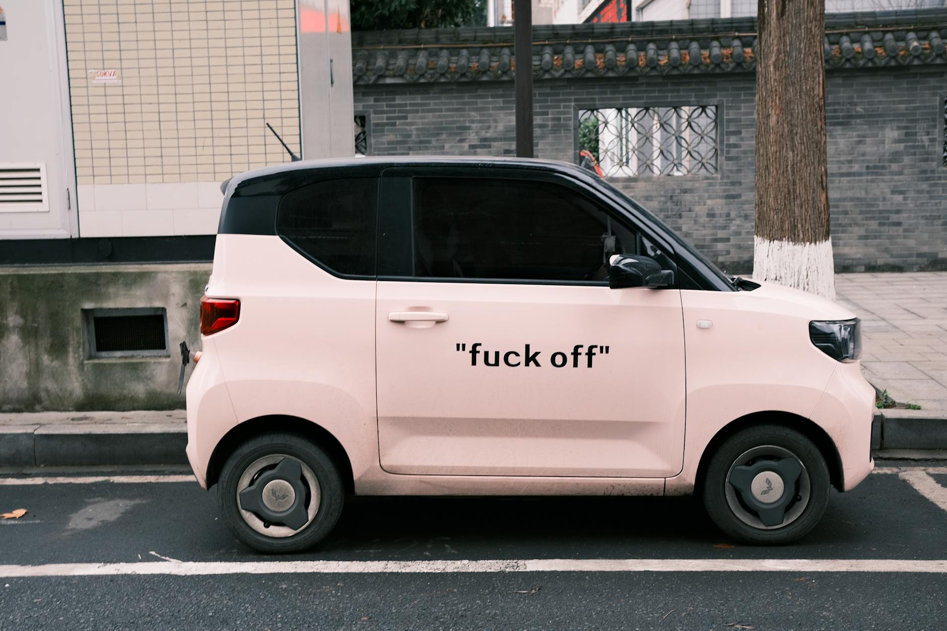 Ilustrasi: Side view of a pink compact electric vehicle parked on an urban street with bold text on the side.